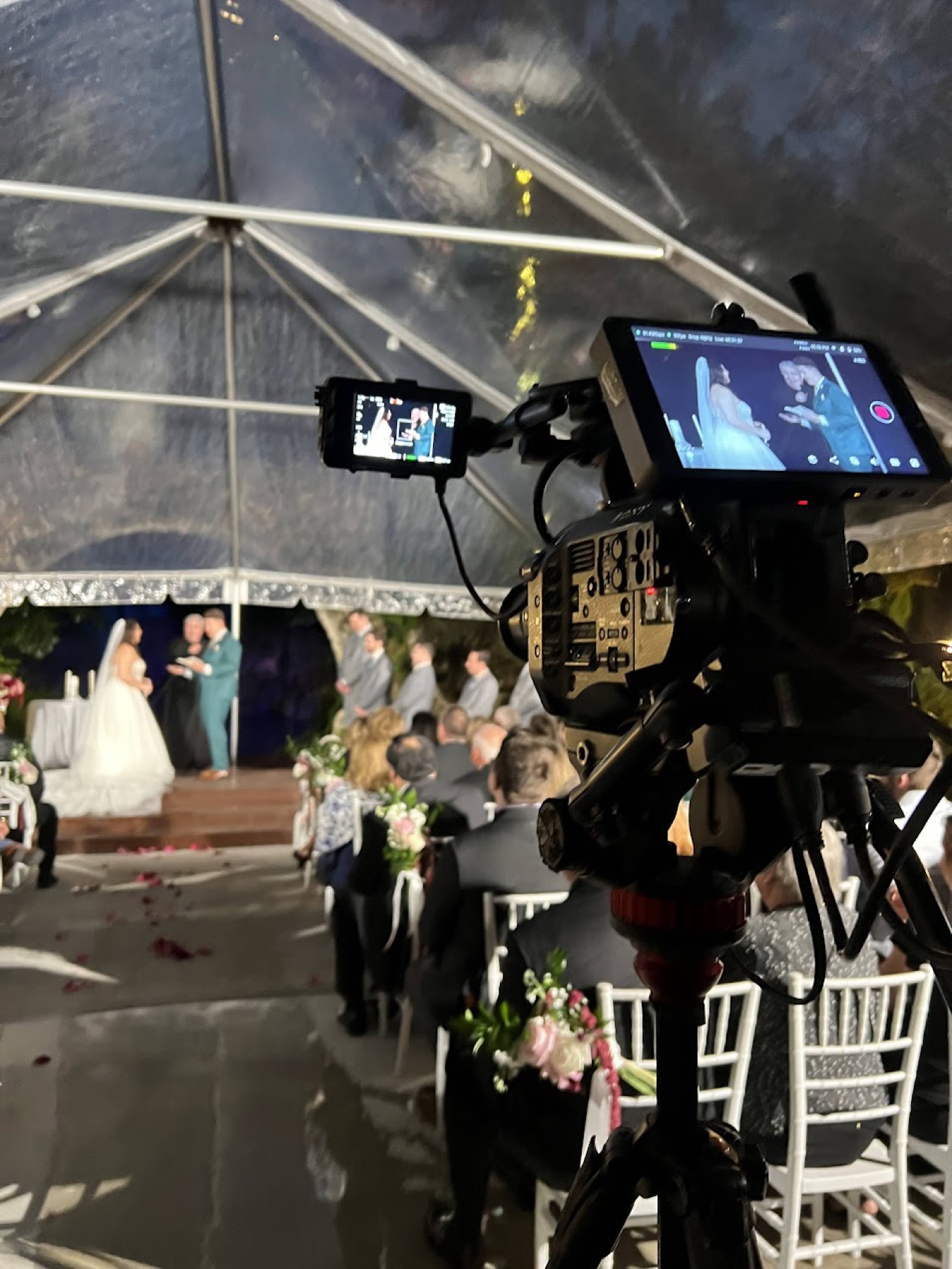 Two production monitors recording a wedding ceremony under a clear tent while it rains