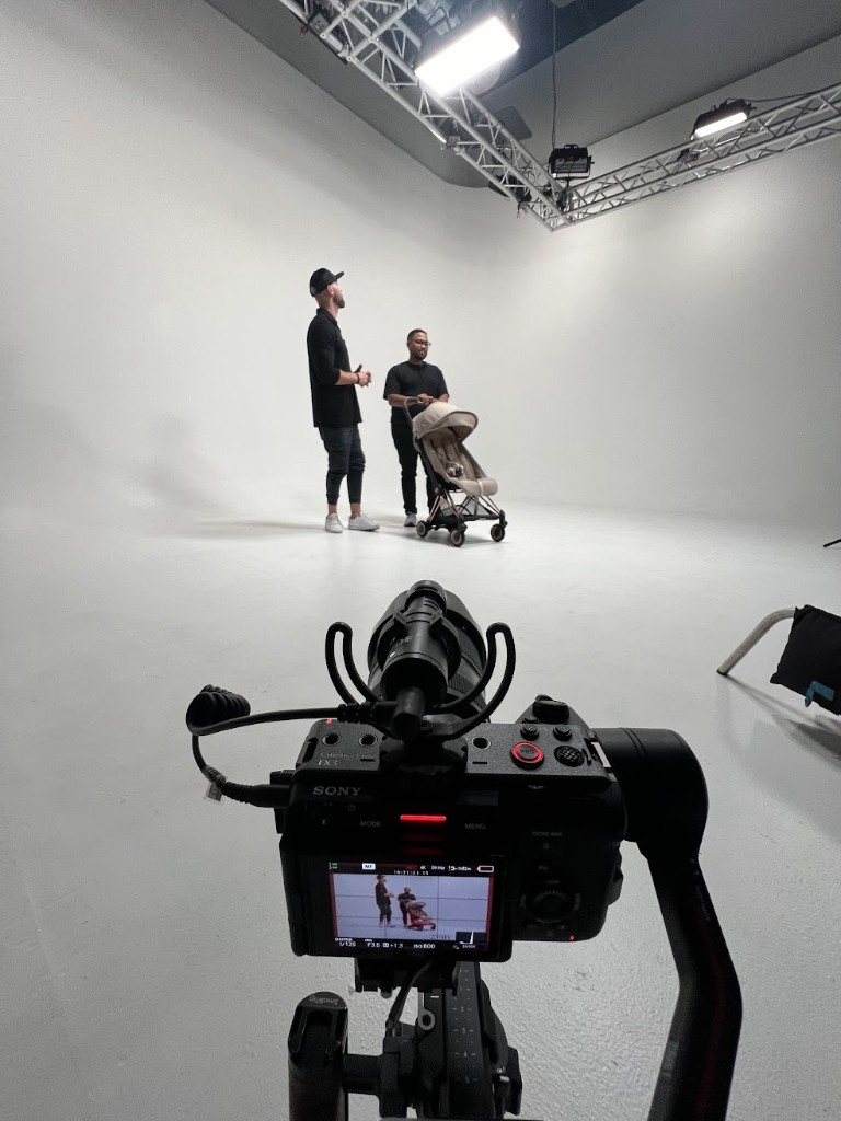 Behind the scenes in a white cyclorama studio: Sony FX3 on a gimbal filming two presenters with a baby stroller product, overhead LED panels